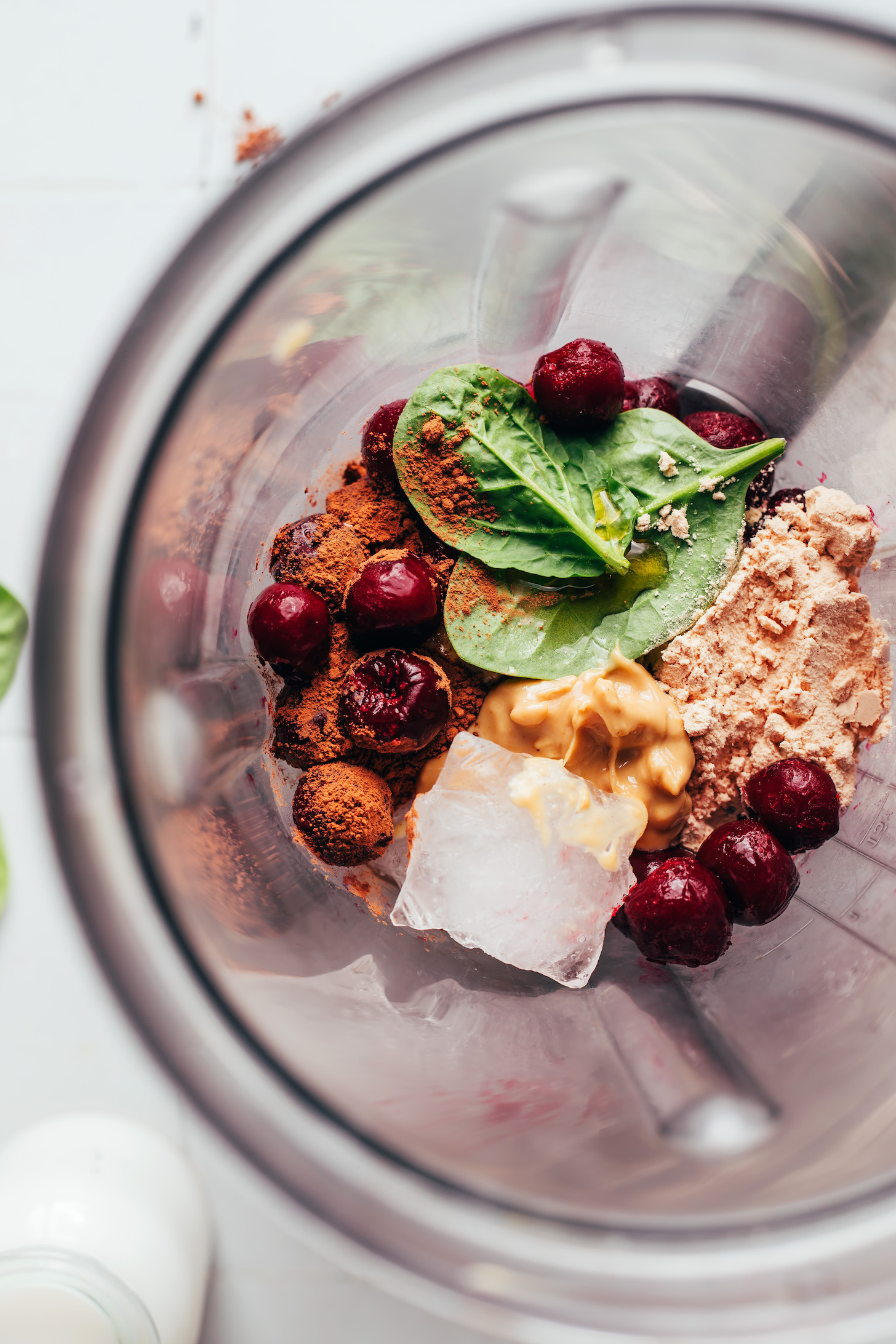 Blender with frozen cherries, ice, cashew butter, protein powder, cocoa powder, and baby spinach